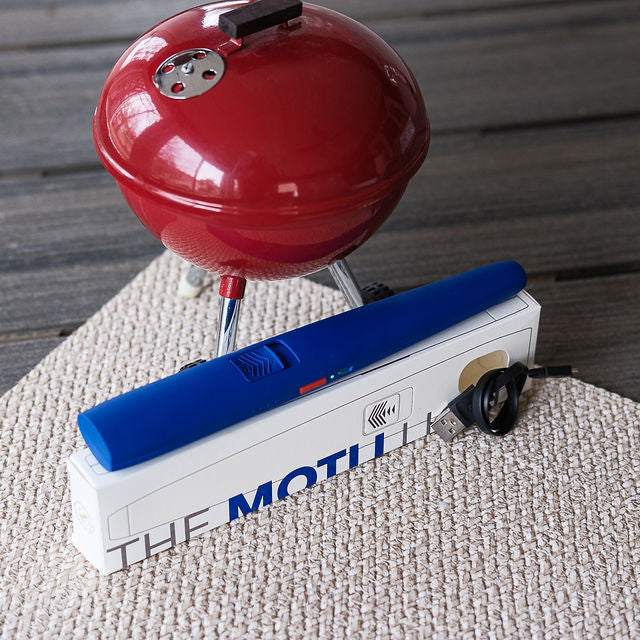 Red portable grill on a white box with 'The Motu' branding, placed on a textured surface.