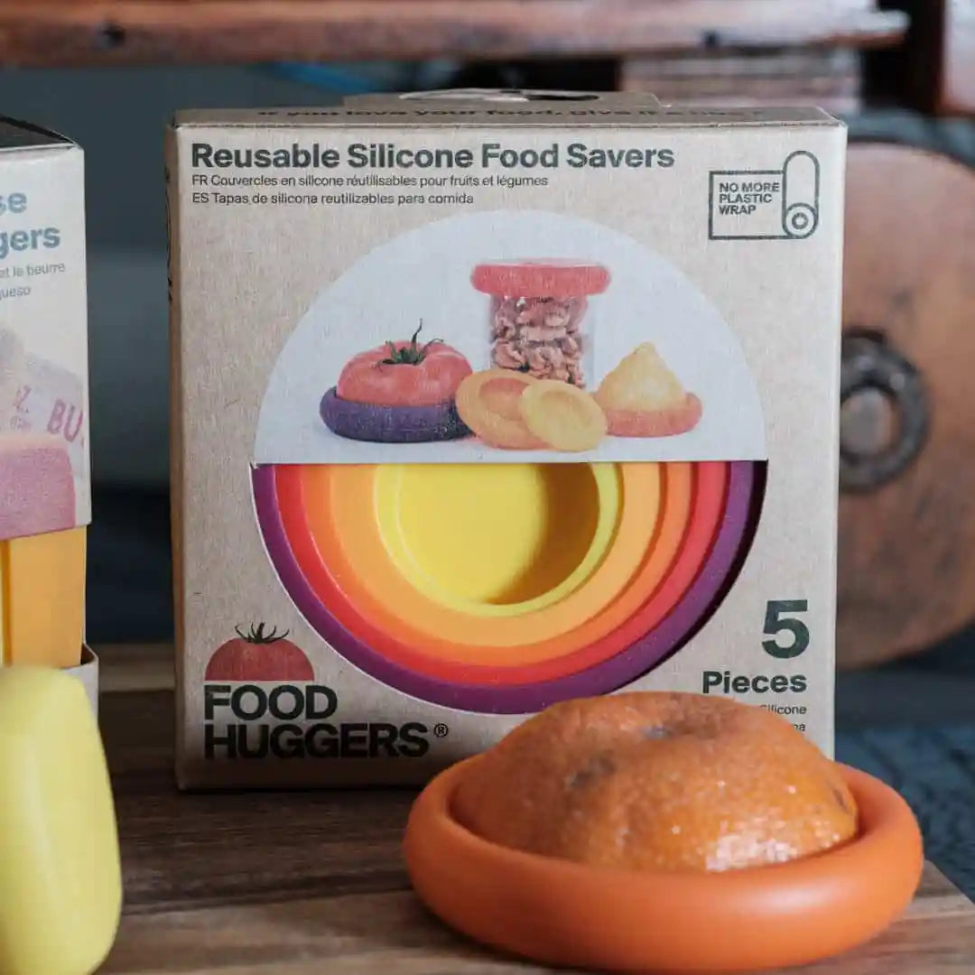 Box of Food Huggers reusable silicone food savers with an orange product on a wooden surface.