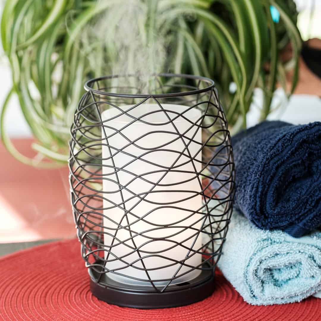 Decorative candle holder with a white candle on a red surface, surrounded by blue and green towels and a plant.