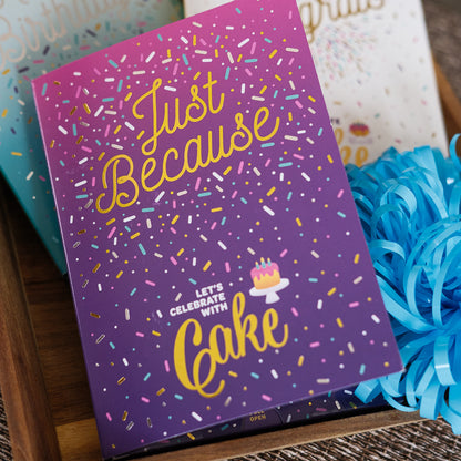 Purple card with colorful text and sprinkles, featuring a cake illustration, on a wooden surface with blue flowers.